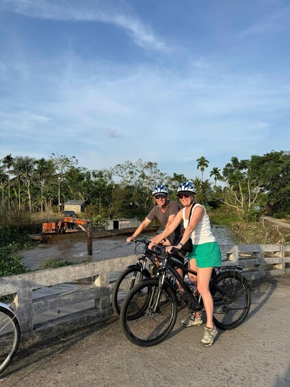 Two people on bicycles near a waterway with a bridge and greenery in the background. 3