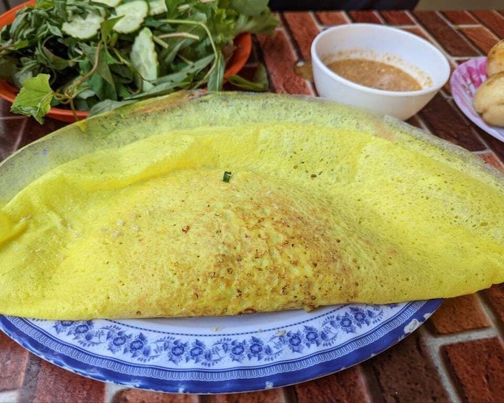 A folded crepe on a blue and white plate, with a bowl of sauce and fresh greens in the background. 3