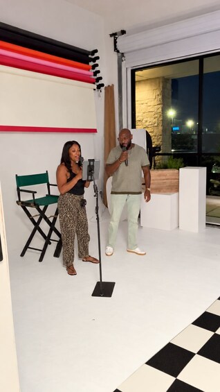 Two people speaking into microphones on a stage with a checkered floor and a large window in the background. 4