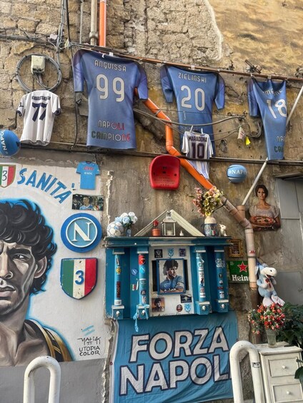 A wall decorated with Napoli football jerseys, a mural of a player, and a banner reading 'FORZA NAPOLI'. 2