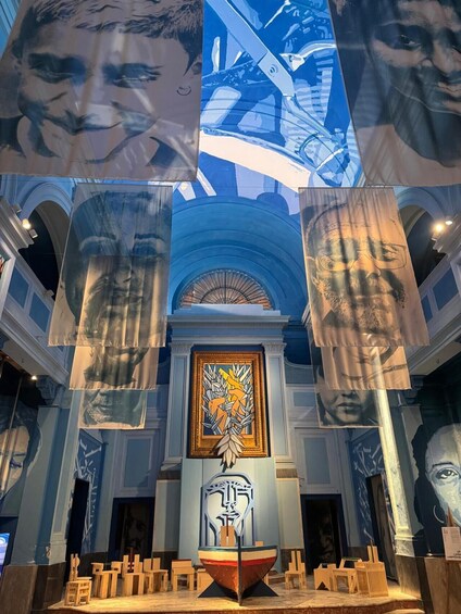 An interior with a central stained glass window, wooden chairs, and hanging banners with portraits. 1