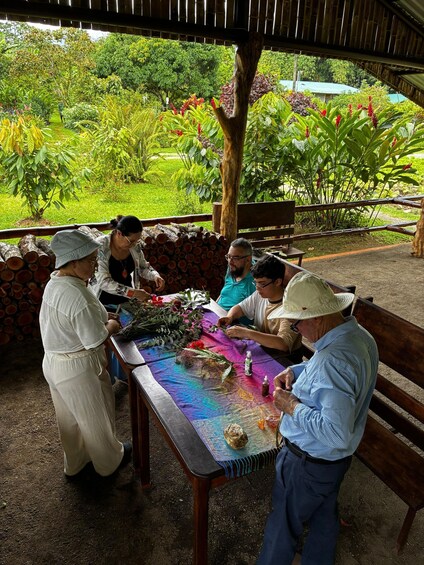 A group of people engaged in a craft activity outdoors, surrounded by lush greenery and a bamboo structure. 1