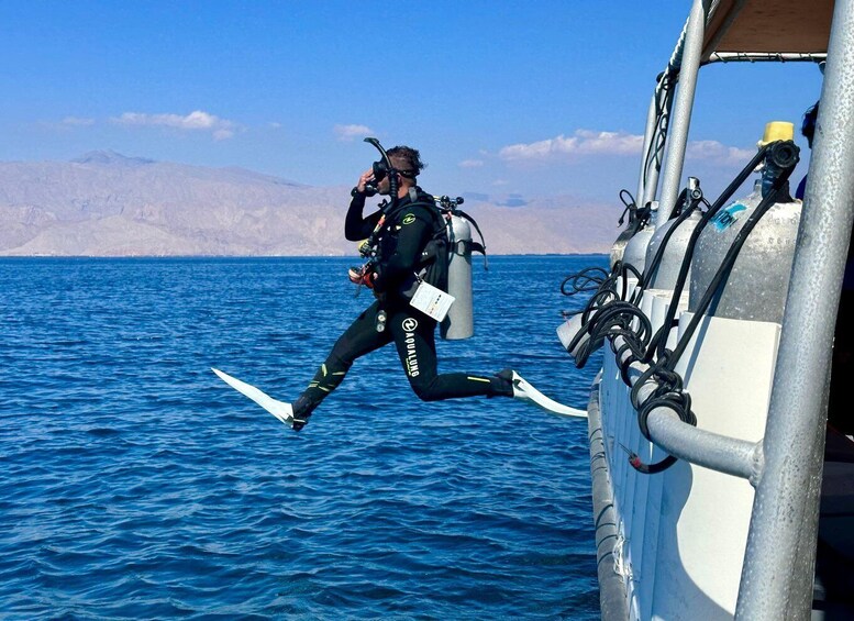A diver in a black wetsuit with fins and a breathing apparatus, preparing to enter the water from a boat. 1