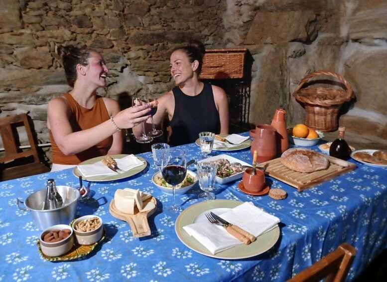 Two people toasting with wine glasses at a table with a blue floral tablecloth, surrounded by various dishes and a rustic stone wall. 1