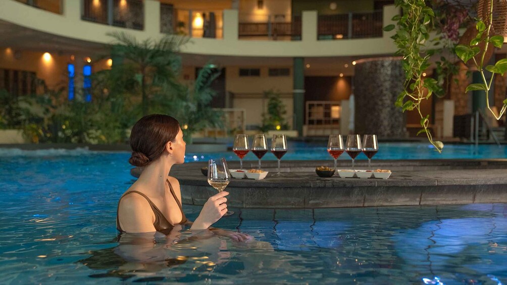 A woman in a swimming pool holding a glass of wine, with several glasses and snacks on the poolside. 2