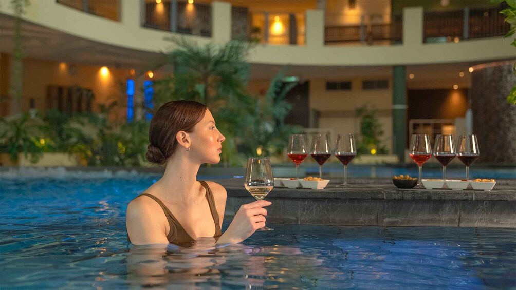 A person in a pool holding a wine glass, with several other wine glasses and snacks on a table in the background. 4