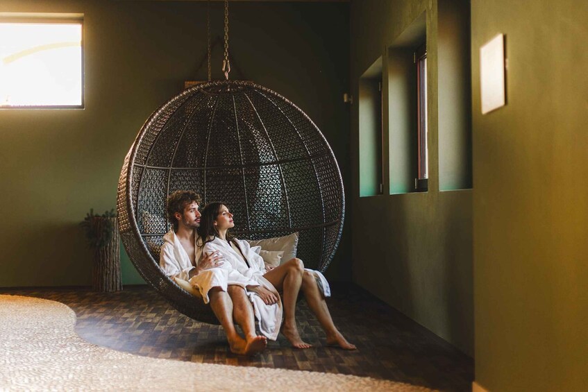 A couple sitting in a woven hanging chair in a room with a wooden floor and a window. 5