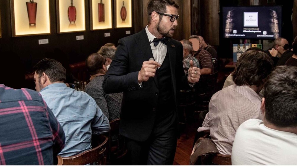 A man in a black suit walking through a crowded bar with people seated. 2