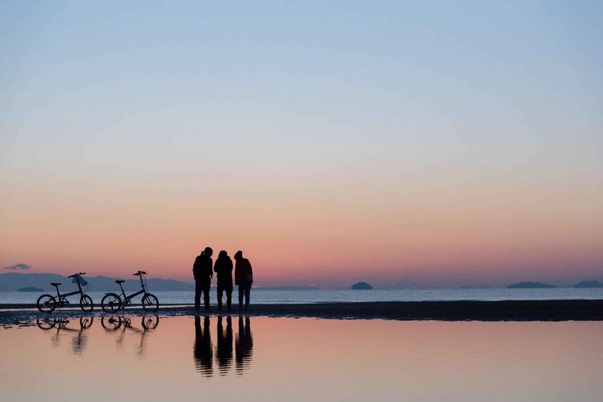 Three people standing on a beach with bicycles, reflecting on the water during sunset. 1