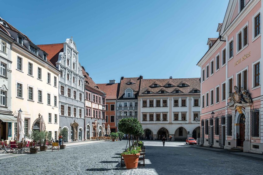 A cobblestone square with historic buildings, a central tree, and outdoor seating. 1