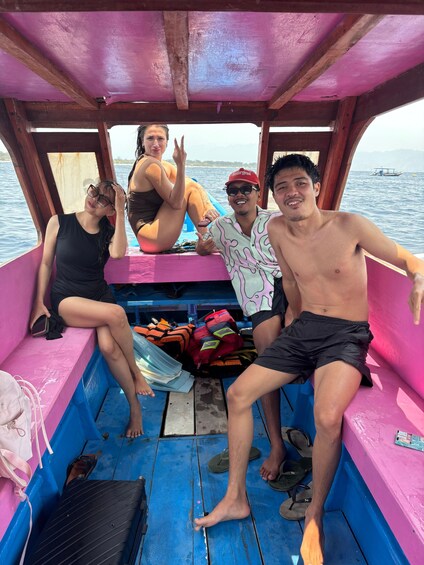 A group of people on a boat with a pink interior, some sitting and some standing, with a body of water and a boat in the background. 3