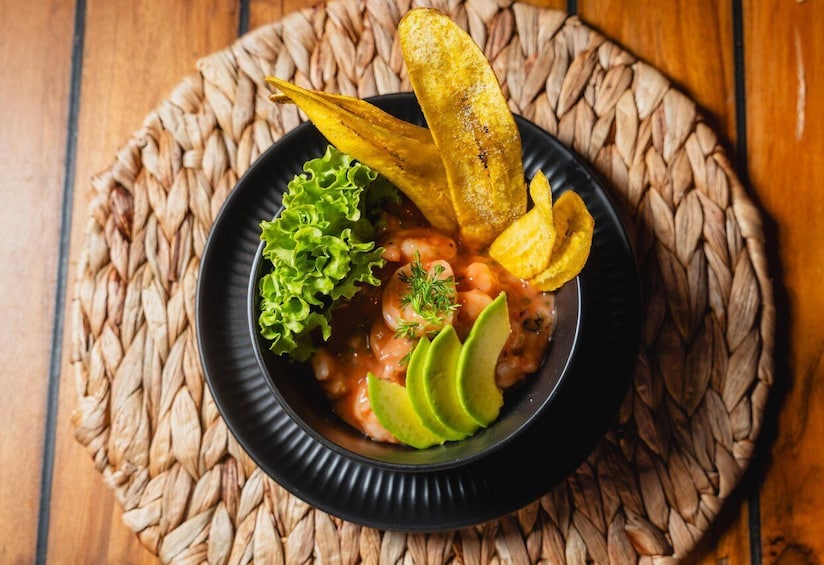 A bowl of food with shrimp, avocado slices, and a side of plantains on a woven mat. 2