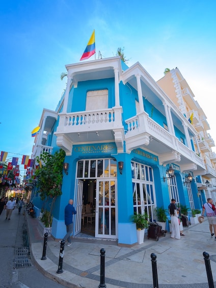A blue and white building with a flag, named 'CENTENARI Restaurant Bar,' and a street with pedestrians. 1