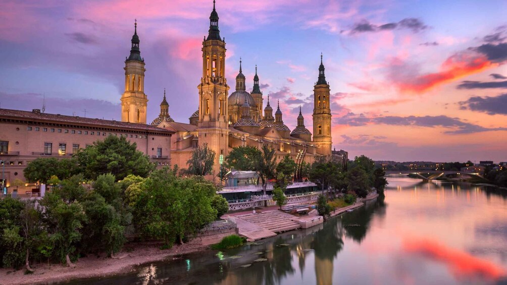 A historic cathedral with multiple spires, a river in the foreground, and a sunset sky. 1