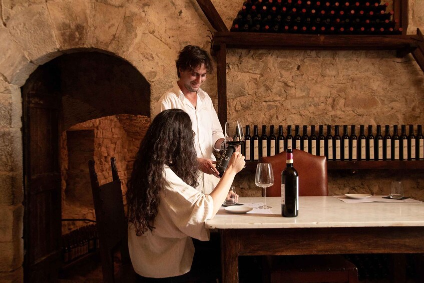 A person pouring wine into a glass at a table in a stone-walled room with a wine rack in the background. 1