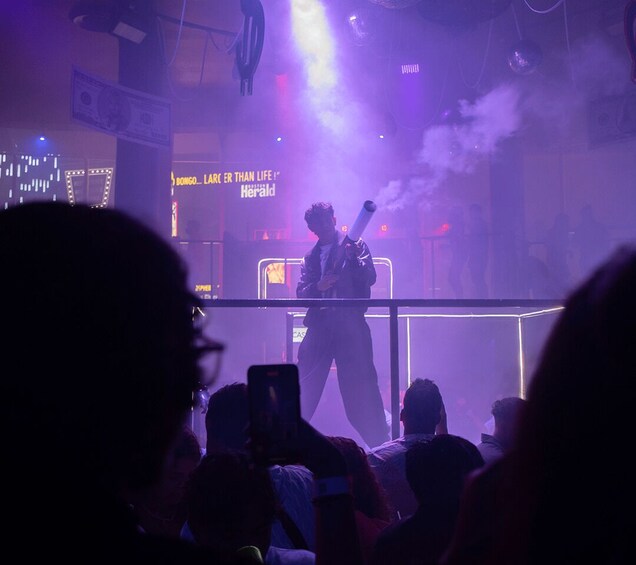 A person performing on stage with a crowd in front, under purple stage lighting. 1