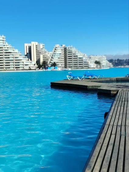 A wooden pier extending into a clear blue pool with lounge chairs and a cityscape in the background. 1