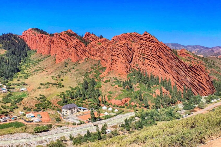 A mountainous landscape with red rock formations, a small settlement with houses and a building, and a winding road. 1