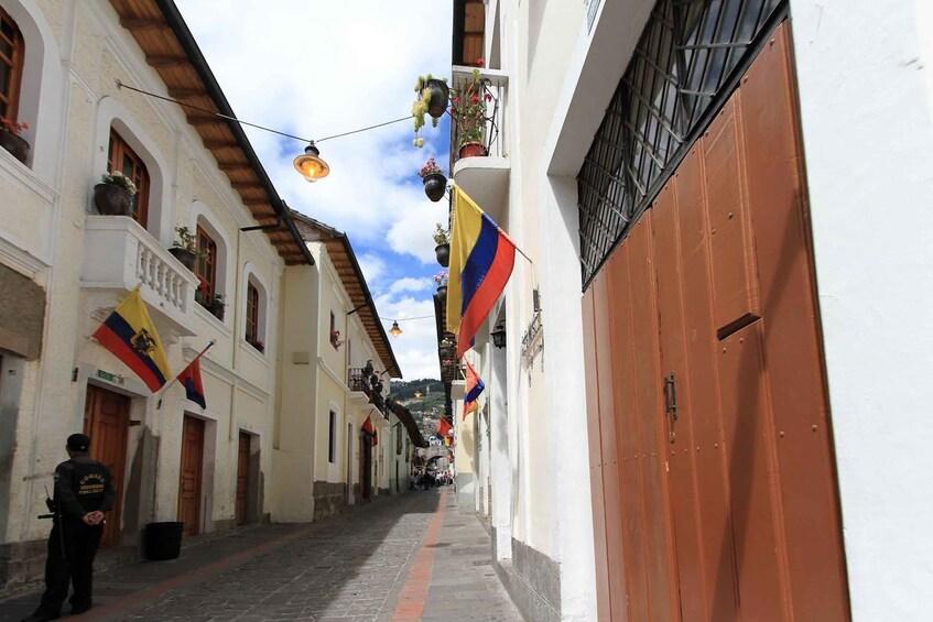 A narrow street with white buildings, flags, and a person standing by a door. 4
