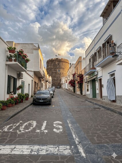 A cobblestone street lined with buildings, a stone tower, and parked cars. 1