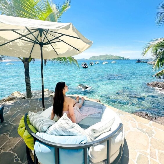 A person relaxing on a stone deck with a pool chair, overlooking a clear blue sea with boats and palm trees. 5