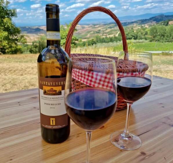 A bottle of Santavenere wine, two glasses of red wine, and a wicker basket on a wooden table with a scenic view in the background. 5