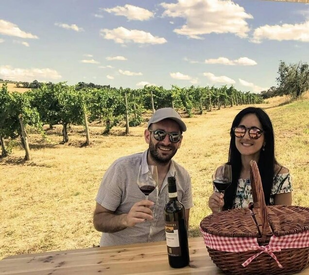 Two people sitting at a table outdoors with a bottle of wine and a basket. 1