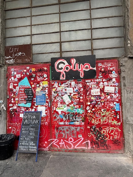 A red door covered in stickers and graffiti, with a neon sign above and a chalkboard sign to the left. 2