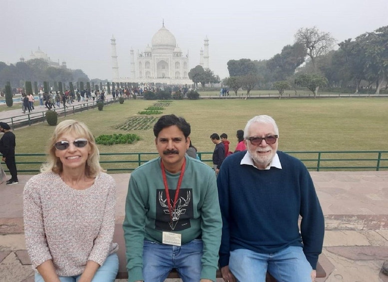 Three people seated in front of the Taj Mahal with a park and visitors in the background. 3