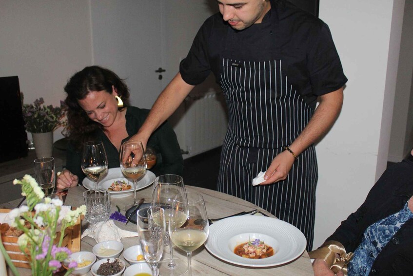 A person in a striped apron serving food at a table with wine glasses and dishes. 5