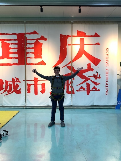 A person in a harness standing in front of a wall with large red Chinese characters and the words 'SKYDIVE CHONGQING' on it. 3