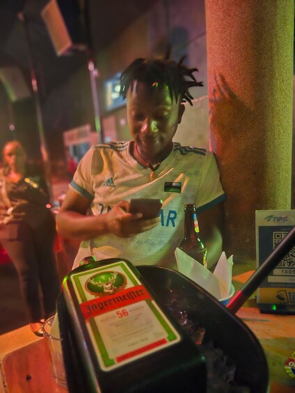 Une personne portant un maillot de sport orné d’un logo, tenant un téléphone intelligent et une bouteille de Jagermeister posée sur une table. 3