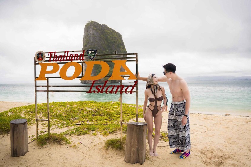 A wooden sign with 'Thailand Poda Island' written on it, a man and woman standing in front of it, and a large rock formation in the background. 1