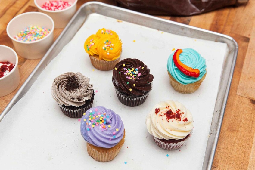 Six cupcakes on a baking tray with various frosting colors and sprinkles. 5
