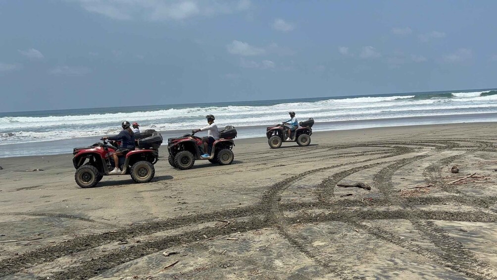 Three people riding ATVs on a sandy beach with ocean waves in the background. 3