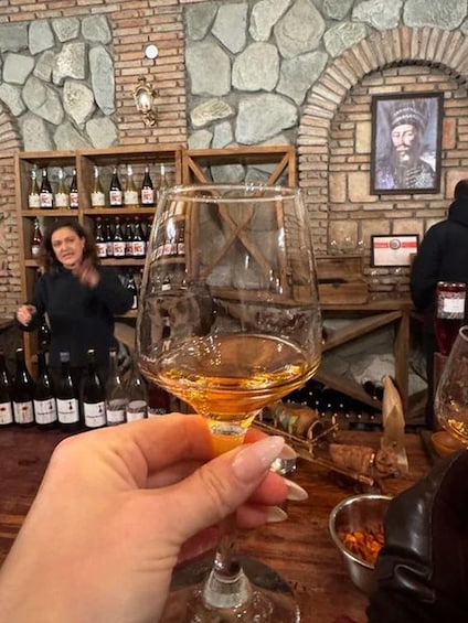 A hand holding a wine glass with a golden liquid, a stone wall, and a person in the background. 5