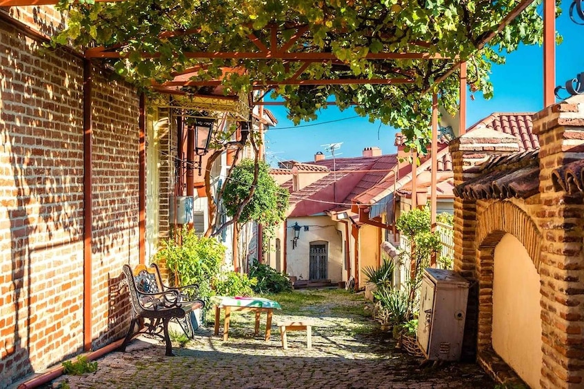A cobblestone alley with a bench, a table, and a pergola covered in vines. 1