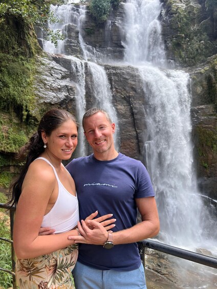 A couple standing in front of a waterfall with lush greenery and rocky terrain. 1