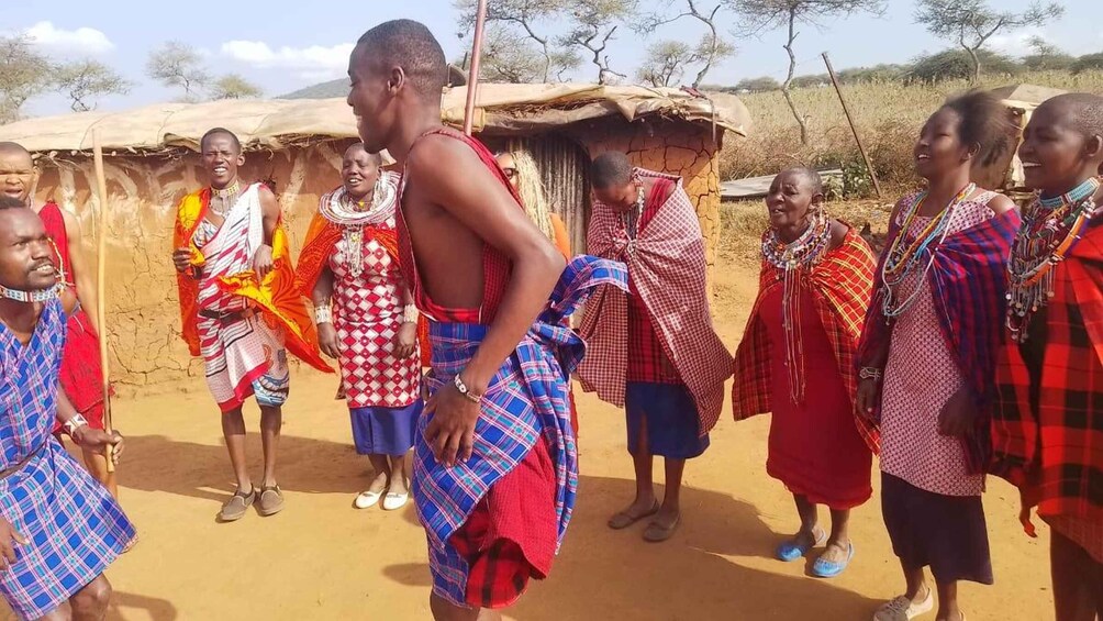 A group of people in traditional attire standing in a rural setting with huts in the background. 1