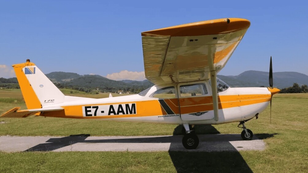 A small propeller airplane with registration E7-AAM parked on a grassy field with mountains in the background. 2
