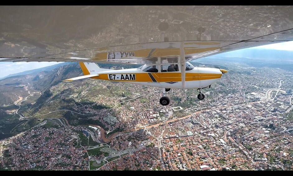 A yellow and white aircraft with registration E7-AAM flying over a densely populated city with visible roads and buildings. 1