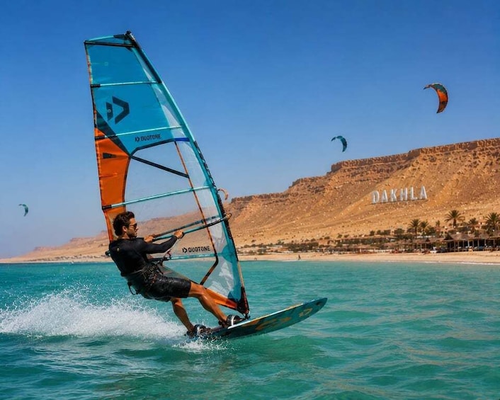 A person windsurfing on turquoise water with a clear sky and a dune in the background. 1