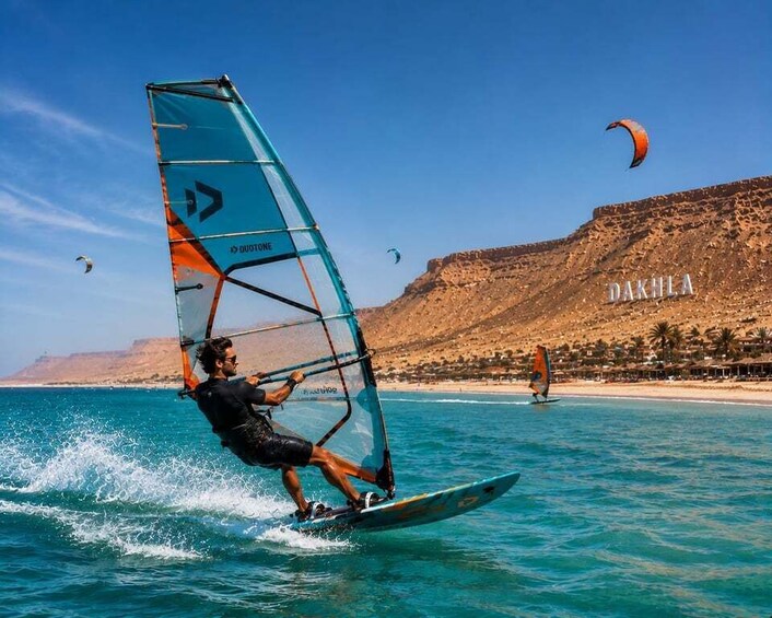 A person windsurfing on turquoise waters with a clear sky and a mountainous backdrop. 3