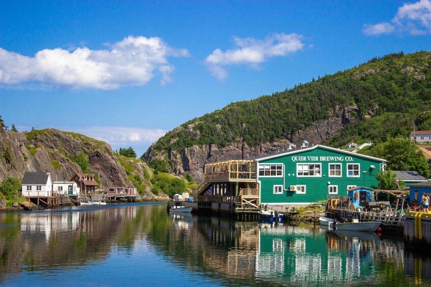 A green building labeled 'QUIDI VIDI BREWING CO.' by a calm lake with boats and a forested hill in the background. 3