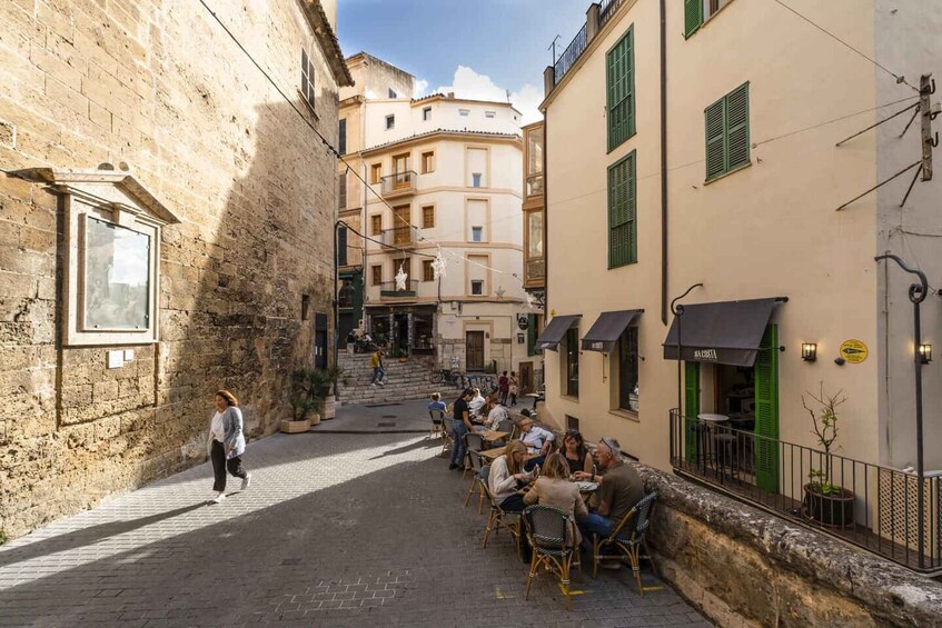 A narrow street with people sitting at outdoor cafes, a building with a balcony, and a staircase leading to a higher floor. 2
