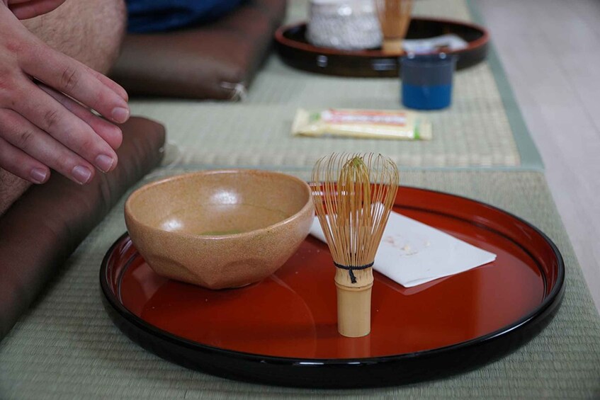 A wooden bowl and whisk on a red tray, with a green mat and a person's hand in the background. 1