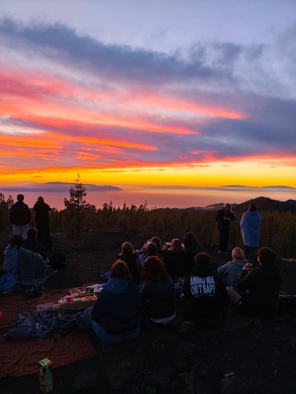 A group of people sitting on a blanket at sunset, overlooking a forested landscape. 1