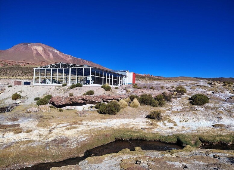 A modern glass structure with a red section, set in a dry, rocky landscape with sparse vegetation and a mountain in the background. 1