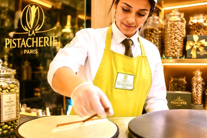A person in a yellow apron working with a large black pan, with jars of pistachios and a sign reading 'La Pistacherie Paris' in the background. 3