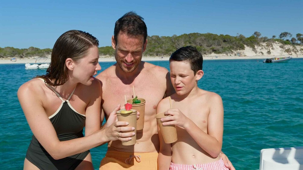 Three people on a boat with drinks, a sandy beach, and clear blue water in the background. 4
